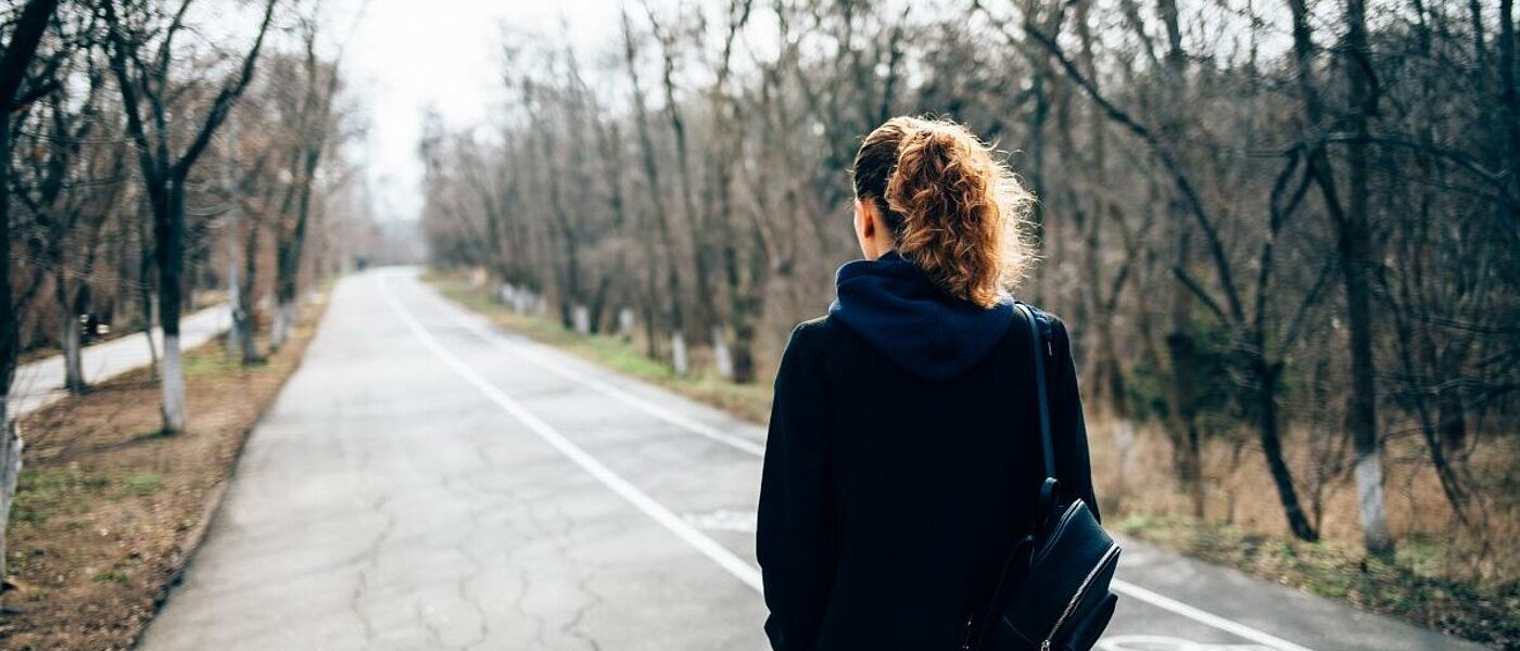 Eine junge Frau mit hellen lockigen Haaren und einer Tasche über den Schultern, läuft eine leere Straße entlang und ist von hinten zu sehen. 