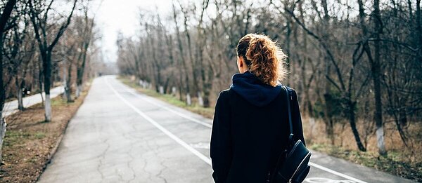 Eine junge Frau mit hellen lockigen Haaren und einer Tasche über den Schultern, läuft eine leere Straße entlang und ist von hinten zu sehen. 