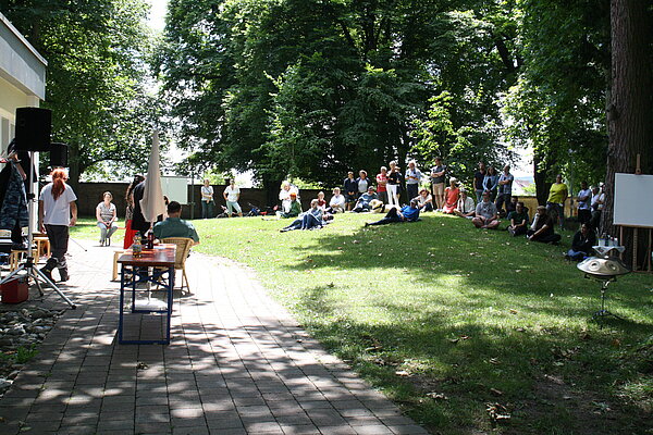 Auf einer Wiese unter Bäumen sitzen und liegen mehrere Personen. Sie schauen alle nach links in die selbe Richtung, wo mehrere Personen ein Theaterstück aufführen.