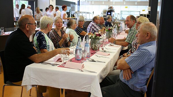 An einem langen festlich gedeckten Tisch sitzen mehrere ältere Männer und Frauen zusammen. Sie unterhalten sich untereinander. Im Hintergrund sind Küchenkräfte und Bedienungen in weißer Arbeitskleidung zu sehen.
