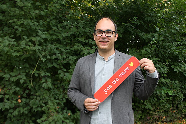 Ein Mann mit Brille steht vor einem Strauch und hält ein auffällig rotes Schild in den Händen, darauf steht mit weißer Schrift "yes we care" und danach folgt ein gelbes Herz.