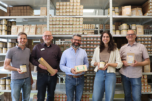 Vor einem großen, hohem Regal mit lauter Gewürzpackungen stehen fünf Personen nebeneinander. Sie alle halten eine Gewürzpackung in den Händen und schauen freundlich in die Kamera.