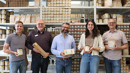 Vor einem großen, hohem Regal mit lauter Gewürzpackungen stehen fünf Personen nebeneinander. Sie alle halten eine Gewürzpackung in den Händen und schauen freundlich in die Kamera.