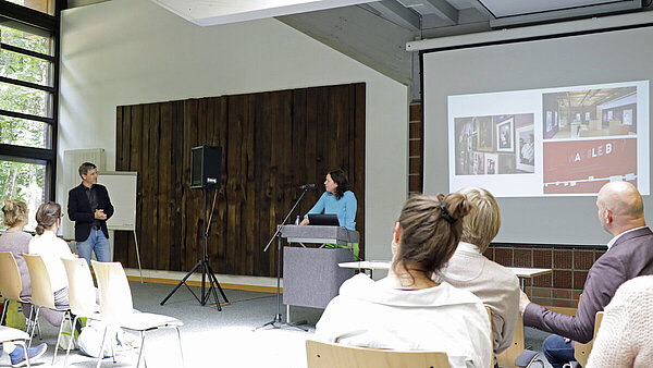Moderator und Referentin tauschen sich aus, Zuhörerinnen und Zuhörer im Vordergrund, im Hintergrund rechts ist eine Bildschirmpräsentation zu sehen.
