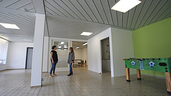 Zwei Mitarbeiterinnen des ZfP Südwürttemberg stehen in einem großen Raum, sie sind einander zugewandt und unterhalten sich. Rechts im Bild steht ein Tischkicker, daneben befindet sich der Zugang zu einer Küchenzeile. Hinter den beiden Frauen ist eine geöffnete Schiebetür, die den Blick freigibt in einen weiteren Raum, in dem zwei große Tische sowie Stühle stehen.