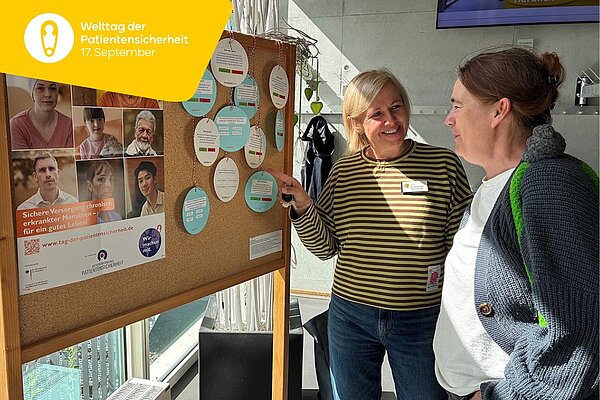 Zwei Frauen stehen, sich unterhaltend, neben einer Stellwand, auf der ein Plakat und ein Quiz zur Patientensicherheit angepinnt sind.