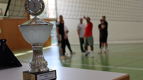 Man sieht einen großen goldenen Pokal im Vordergrund, im Hintergrund ist ein Volleyballfeld erkennbar auf dem 4 Personen beisammen stehen.