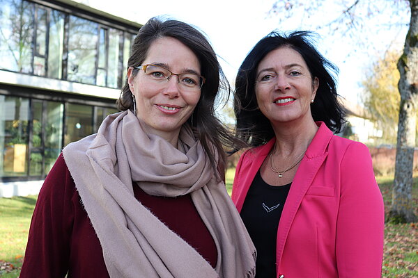 Zwei Frauen lachen in die Kamera. Links, braunhaarig mit Brille und rosanem Schal und rechts schwarze Harre, pinker Lippenstift und pinker Blazer mit schwarzem Shirt.