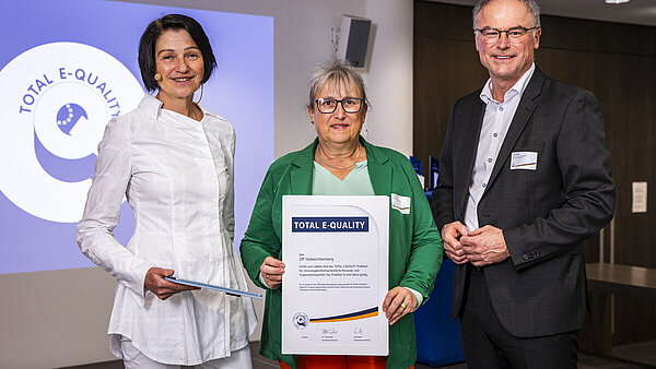 In einem Veranstaltungssaal stehen zwei Frauen und ein Mann vor dem Logo des Vereins Total E-Quality, die Frau in der Mitte hat eine Urkunde in der Hand.