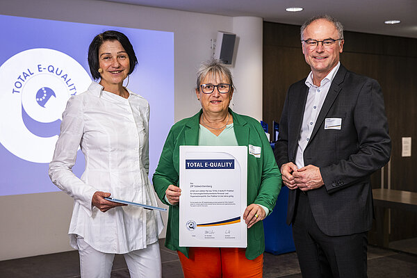 In einem Veranstaltungssaal stehen zwei Frauen und ein Mann vor dem Logo des Vereins Total E-Quality, die Frau in der Mitte hat eine Urkunde in der Hand.