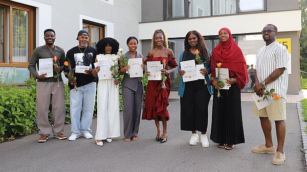 Acht junge Erwachsene verschiedener Nationalitäten stehen vor einem Klinikgebäude und halten ihre Abschlussurkunden in den Händen.