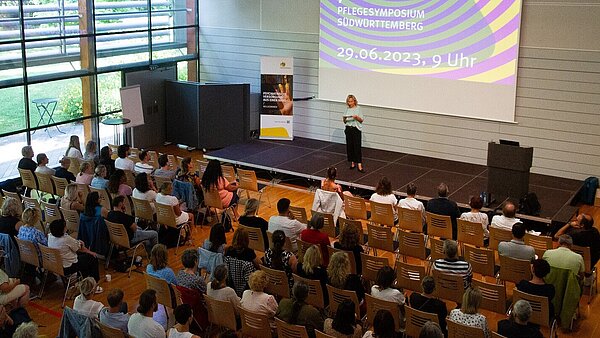 In mehreren Reihen sitzen Zuhörer:innen und schauen auf eine Bühne, auf der eine Frau moderiert. 