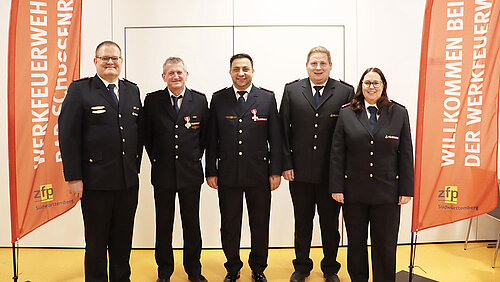Vier Männer und eine Frau in Feuerwehr-Ausgehuniform, flankiert von zwei roten Fahnen mit der Aufschrift Werkfeuerwehr Bad Schussenried.