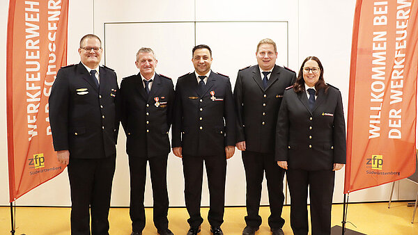 Vier Männer und eine Frau in Feuerwehr-Ausgehuniform, flankiert von zwei roten Fahnen mit der Aufschrift Werkfeuerwehr Bad Schussenried.