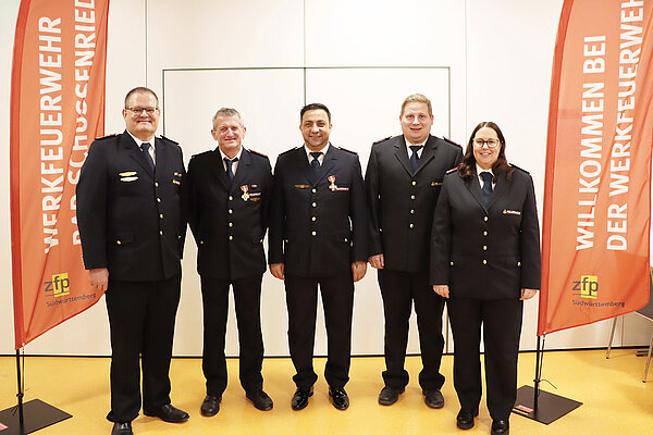 Vier Männer und eine Frau in Feuerwehr-Ausgehuniform, flankiert von zwei roten Fahnen mit der Aufschrift Werkfeuerwehr Bad Schussenried.