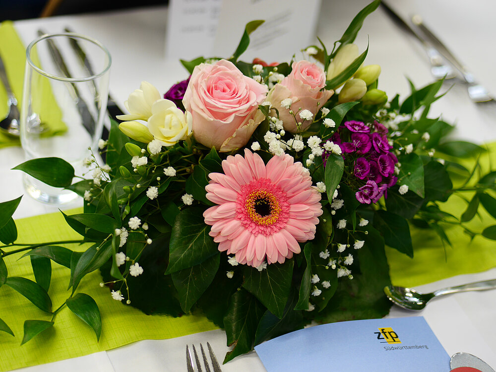 Nahaufnahme: Auf einem gedeckten Tisch mit weißer Tischdecke und hellgrünem Tischläufer steht ein Blumengesteck. In dem Gesteck sind grüne Blätter, kleine weiße Blüten, rosafarbene Rosenblüten, kleine lila Blumen und eine große rosafarbene Gerbera-Blüte. An dem Tischplatz unten in der Mitte liegt zwischen dem Besteck eine Broschüre mit ZfP-Logo oben in der Ecke.