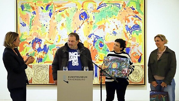 Drei Frauen und ein Mann stehen vor einem farbenfrohen Gemälde in einem Museum und sprechen an einem Rednerpult miteinander. 