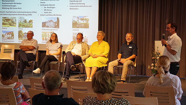 Zwei Frauen und drei Männer sitzen auf einer Bühne, im Vordergrund steht ein Mann mit Mikrofon. 