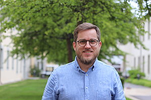 Ein jüngerer Mann mit kurzen, hellbraunen Haaren, Brille und einem hellblauen Hemd. 