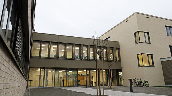 Ein geradliniger, beige-brauner Gebäudekomplex mit hell erleuchteten Fenstern, im Vordergrund des Bildes ist etwas Hof zu sehen, in der Mitte der Haupteingang mit einer breiten Glasfront und Schiebetüren.