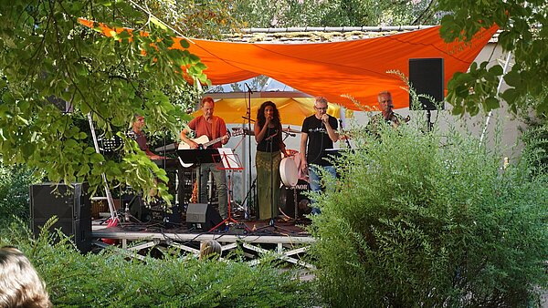 Auf einer Bühne umgeben von viel Grün spielen Musiker unterschiedliche Instrumente. 