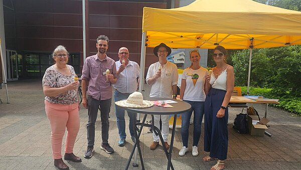 Eine Gruppe Menschen steht vor einem gelben Pavillon, alle halten ein Eis in der Hand. 