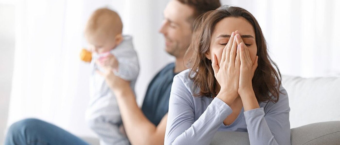 Eine junge, verzweifelt wirkende, Frau sitzt auf einem Sofa und hält die Hände vor ihr Gesicht. Im Hintergrund ist ein lachender Mann zu sehen, der ein Baby hält.