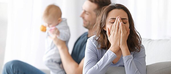 Eine junge, verzweifelt wirkende, Frau sitzt auf einem Sofa und hält die Hände vor ihr Gesicht. Im Hintergrund ist ein lachender Mann zu sehen, der ein Baby hält.