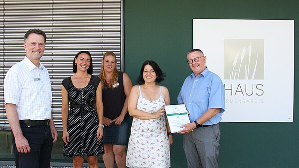 Zertifikatsübergabe im Haus Irmengardis Fünf Personen stehen vor einem grünen Gebäude mit Jalousien an der Fassade. Alle lächeln in die Kamera. Links und rechts steht jeweils ein Mann, in der Mitte stehen drei Frauen. Der Mann ganz rechts hält eine Urkunde in den Händen, die Frau links neben ihm hält die Urkunde ebenfalls. Ganz rechts neben dem Mann ist an der Fassade des Hauses ein weißes Schild mit dem Aufdruck "Haus irmengardis".