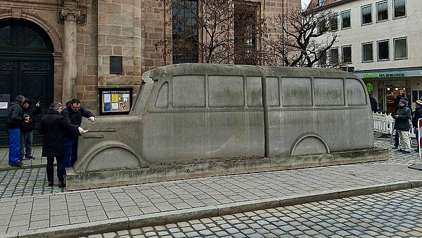 Das Denkmal der grauen Busse, eine mehrere Meter lange Nachbildung eines Omnibusses aus grauem Beton, steht auf gepflasterten Vorplatz einer Kirche. Links und rechts davon stehen Menschen.