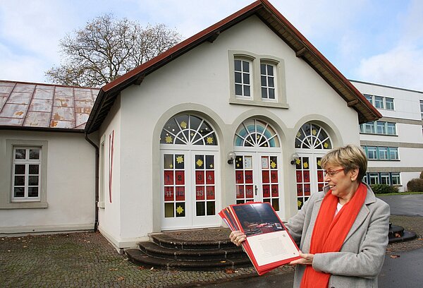 Eine Frau mit rotem Schal steht vor einem Gebäude. Die Fenster sind adventlich geschmückt.