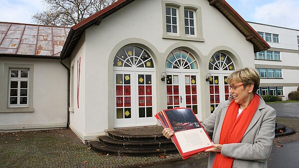 Eine Frau mit rotem Schal steht vor einem Gebäude. Die Fenster sind adventlich geschmückt.