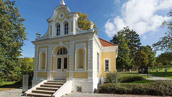Das Württembergische Psychiatriemuseum: Zu sehen ist die Vorderseite mit Treppe und Eingangstüre des barocken Gebäudes, dessen Fassade in weiß und gelb gehalten ist und welches einst eine Friedhofskapelle war. Drumherum ist viel Grün und der blaue, leicht bewölkte Himmel.