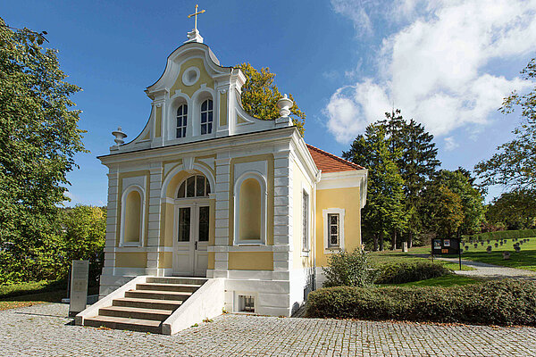 Das Württembergische Psychiatriemuseum: Zu sehen ist die Vorderseite mit Treppe und Eingangstüre des barocken Gebäudes, dessen Fassade in weiß und gelb gehalten ist und welches einst eine Friedhofskapelle war. Drumherum ist viel Grün und der blaue, leicht bewölkte Himmel.