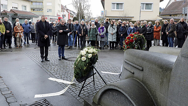 Zwei dunkel gekleidete Männer stehen in stiller Andacht vor einem Denkmal in der Form eines Omnibusses, vor ihnen stehen Gedenkkränze, um sie herum viele ebenfalls anteilnehmende Menschen.