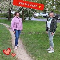 Zwei Frauen stehen nebeneinander auf einer grünen Wiese, am oberen Bildrand ist ein Banner "yes we care" zu sehen. 