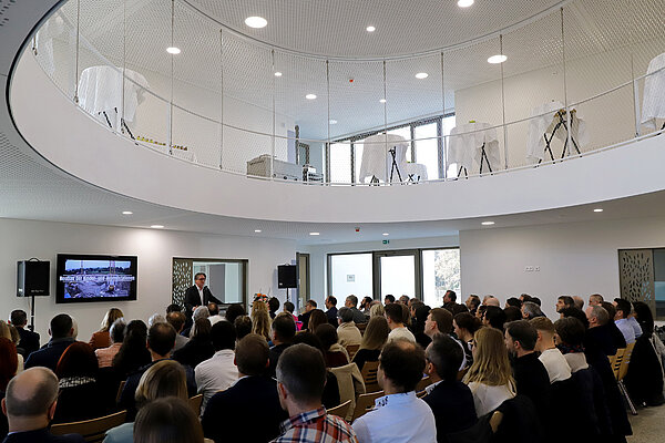 Ein Mann am Rednerpult spricht vor dutzenden sitzenden Zuhörer:innen in einer hellen Eingangshalle, die einen großen ovalen Durchlass in das nächst höhere Stockwerk hat.