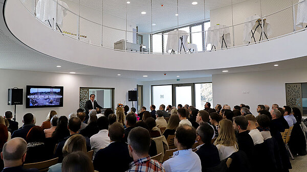 Ein Mann am Rednerpult spricht vor dutzenden sitzenden Zuhörer:innen in einer hellen Eingangshalle, die einen großen ovalen Durchlass in das nächst höhere Stockwerk hat.