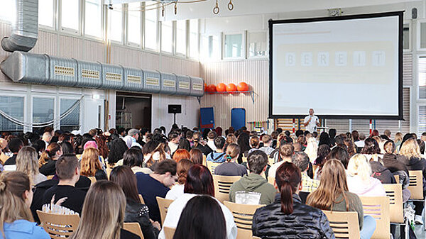 In einer Sporthalle sind viele Stühle aufgestellt, die Halle ist voll besetzt. Der Blick geht vom hinteren Ende über die Köpfe hinweg nach vorne, wo ein Mann am Rednerpult steht.