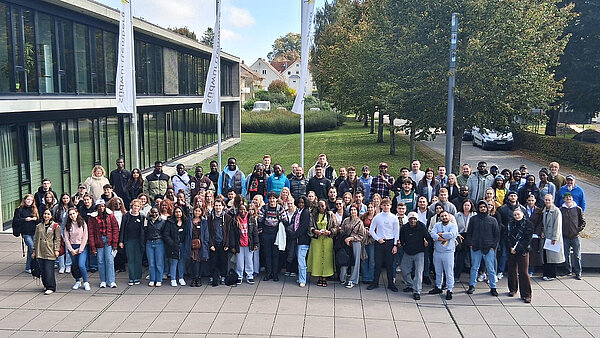 152 junge Frauen und Männer unterschiedlicher Herkunft haben sich vor einem Gebäude aufgestellt und schauen nach oben Richtung Kamera.