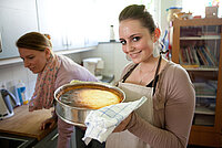 Eine junge Frau hält mit Hilfe eines Tuchs eine Form mit einem Käsekuchen. 