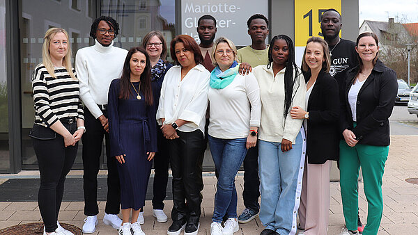Eine Gruppe junger Menschen aus verschiedenen Herkunftsländern steht vor einem Gebäude, welches mit einem gelben Schild mit der Nummer 15 gekennzeichnet ist.