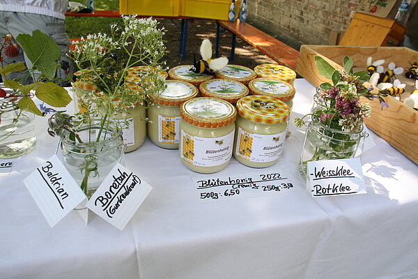 Auf einem Tisch mit weißer Tischdecke stehen mehrere Honiggläser. Daneben liegen verschiedene Blüten- und Pflanzenzweige sowie kleine weiße Schilder mit einer Beschreibung.