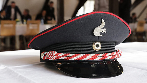 Ein Feuerwehrhut liegt auf einer weißen Tischdecke, im Hintergrund sind Feuerwehrleute sitzend und stehend im Gespräch.