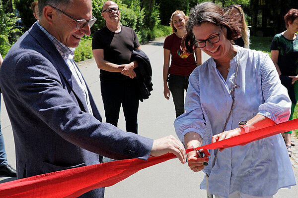 Ein Mann und eine Frau zerschneiden mit einer Schere ein rotes Band, als symbolische Eröffnung des Tabakwanderpfads, im Hintergrund stehen die Teilnehmenden der Premierenwanderung.