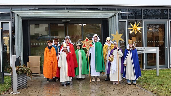 9 Kinder in bunten Gewändern und mit Kronen tragen zwei Sterne mit sich.