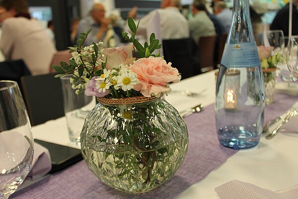 Nahaufnahme eines feierlich gedeckten Tisches. Auf dem Tisch liegen eine weiße Tischdecke und ein lilafarbener Tischläufer. Im Mittelpunkt steht eine runde Glasvase mit einem Strauß aus rosafarbener Rose und weiteren Blumen mit weißen Blüten. Links neben der Vase steht ein rundes Wasserglas, rechts eine Glasflasche Mineralwasser. Im Hintergrund hinter dem Tisch sind sitzende Personen zu sehen.