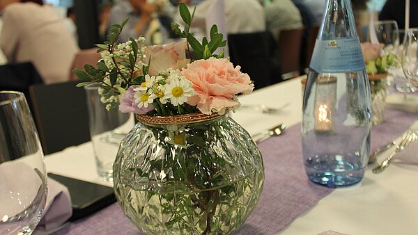 Nahaufnahme eines feierlich gedeckten Tisches. Auf dem Tisch liegen eine weiße Tischdecke und ein lilafarbener Tischläufer. Im Mittelpunkt steht eine runde Glasvase mit einem Strauß aus rosafarbener Rose und weiteren Blumen mit weißen Blüten. Links neben der Vase steht ein rundes Wasserglas, rechts eine Glasflasche Mineralwasser. Im Hintergrund hinter dem Tisch sind sitzende Personen zu sehen.