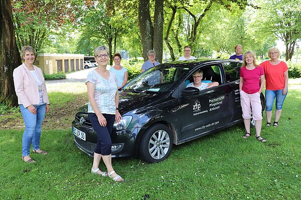 Eine junge Frau sitzt in einem schwarzen Dienstwagen vom Pflegedienst. Sechs Frauen und zwei Männer stehen um das Auto.