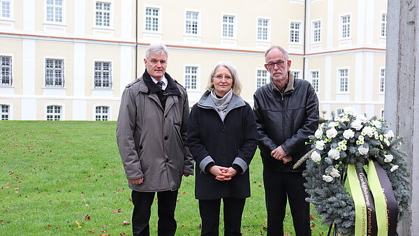 Zwei Männer und eine Frau stehen neben einem Trauerkranz